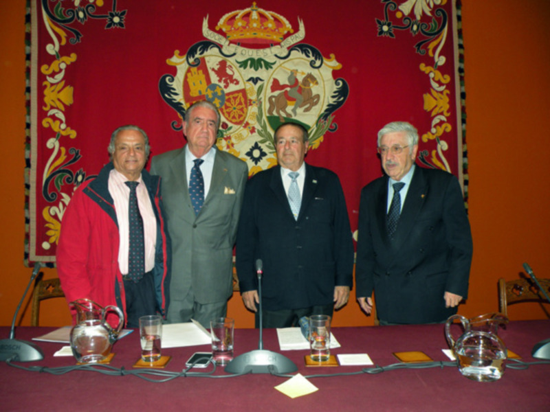 Cuatro hombres en trajes posan frente a un escudo heráldico en una sala formal.