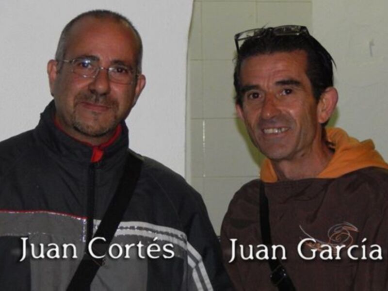Dos hombres sonríen juntos, Juan Cortés y Juan García, en un entorno interior.