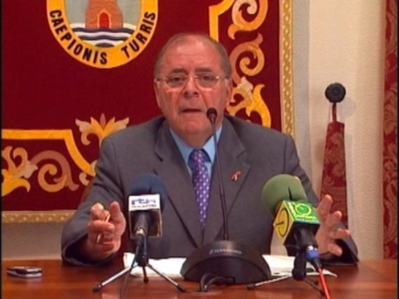 Un hombre en traje y corbata hablando en un micrófono frente a un fondo rojo con un escudo y el texto 'CAEPIONIS TURRIS'.