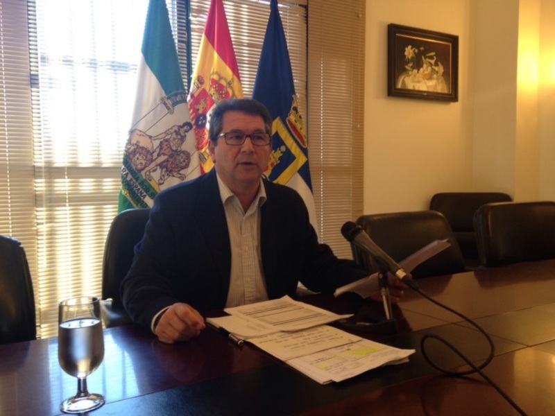Un hombre en una sala de conferencias, frente a una bandera española y un microfono, con documentos en la mesa. La sala tiene una silla en el fondo y una ventana con cortinas. La imagen sugiere una reunión o conferencia formal.