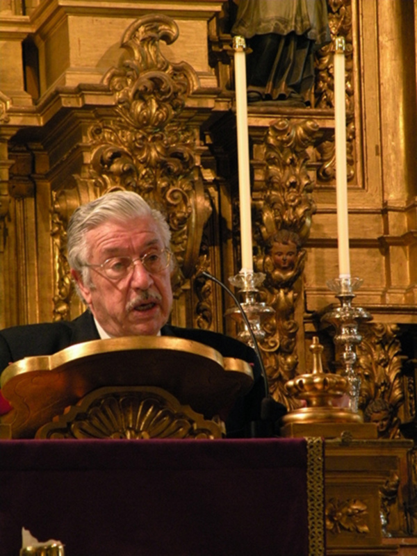 Un hombre habla desde un púlpito ornamental en un entorno de iglesia con velas y detalles dorados.