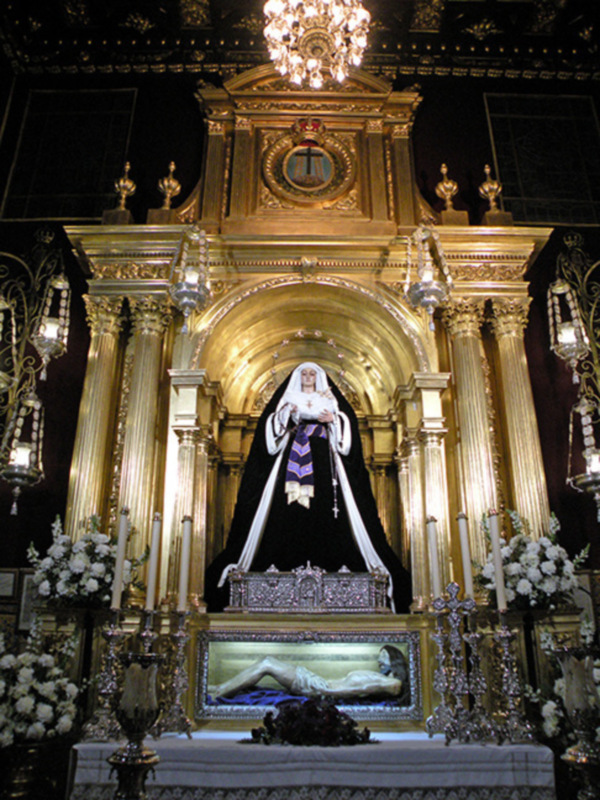 Altar de una iglesia con una imagen de una virgen en el centro, rodeada por flores y adornos dorados, con un reloj en el techo y una lámpara de cristal en la parte superior.