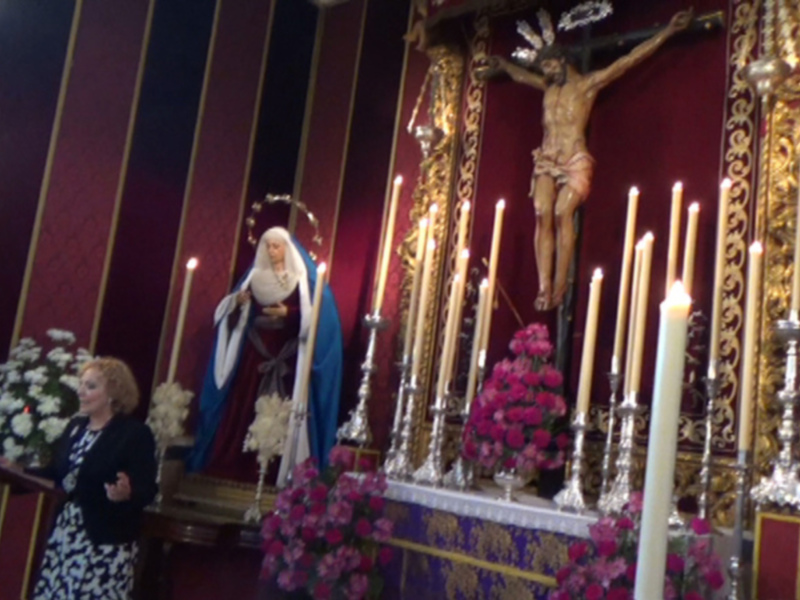 Misa en una iglesia con altar ornado y estatuas religiosas, velas encendidas y flores en honor a la Virgen María.