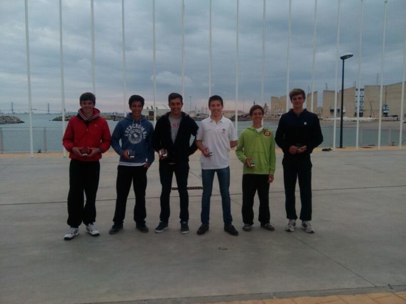 Cuatro jóvenes posan con sus trofeos frente a una bahía con embarcaciones y una estructura metálica en el fondo.
