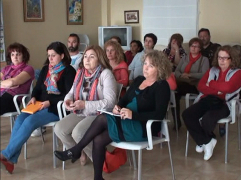 Una sala de conferencias con varias personas sentadas en sillas blancas, algunas sonriendo y otras concentradas. En el fondo hay una televisión y cuadros en la pared.