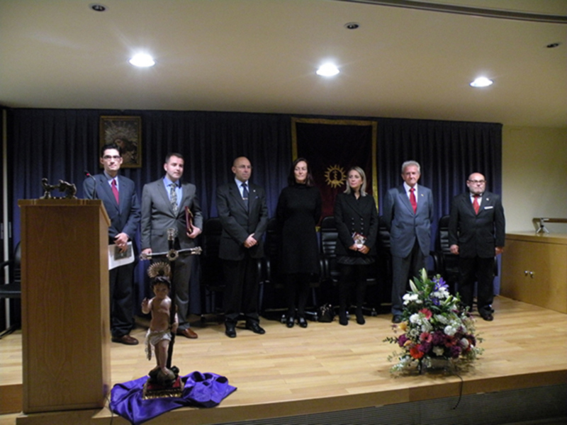 Celebración formal con personas en trajes y una estatua en el escenario.