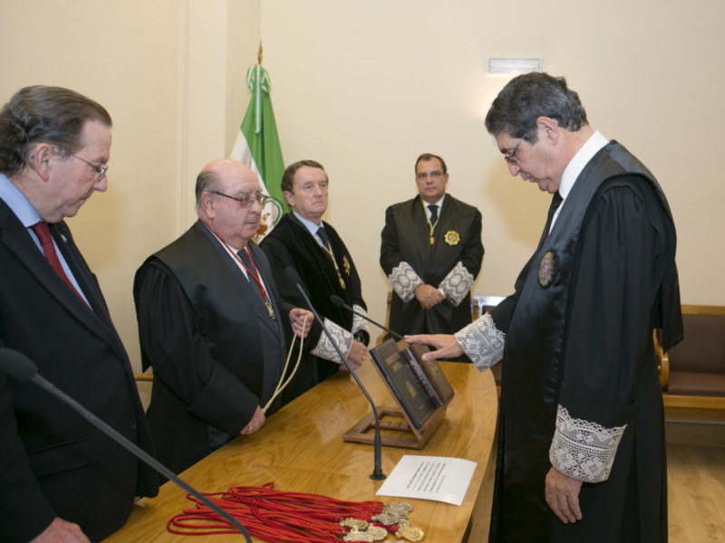 Un hombre en traje negro y corbata de oro se inclina para recibir una medalla de oro de otro hombre en traje negro y corbata de oro, mientras otros hombres observan en un entorno formal con una bandera en el fondo.