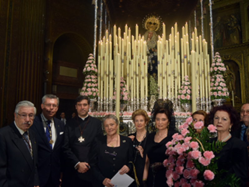 Una procesión religiosa con personas vestidas formalmente y una imagen de la Virgen en el centro.