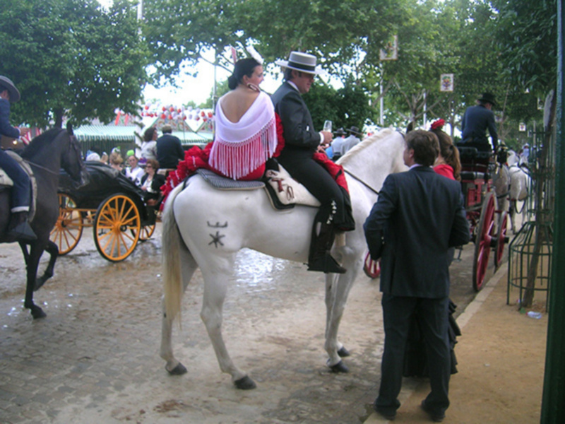 Un par de personas montando en un caballo blanco, con un hombre vestido formalmente y una mujer con un vestido largo y una capa roja, en un ambiente festivo con banderas y carros de caballos en el fondo.