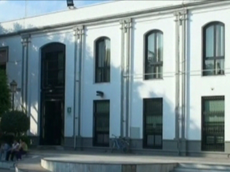 Edificio blanco con ventanas arqueadas y puerta principal, ubicado en un entorno urbano con árboles y personas sentadas en el patio.