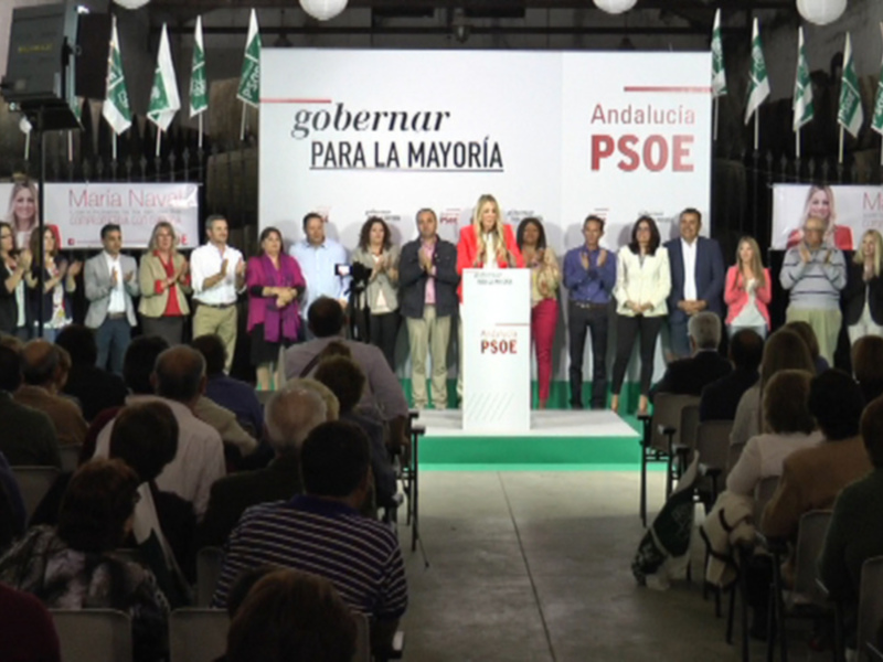 PSOE celebrando en un evento público, con varios miembros del partido saludando a la audiencia. El escenario está decorado con banderas del partido y carteles con mensajes políticos. La audiencia está sentada en filas, observando a los parlamentarios.