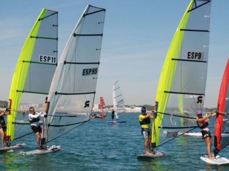 Gente practicando windsurf en el mar con vela amarilla y roja, en un día soleado.
