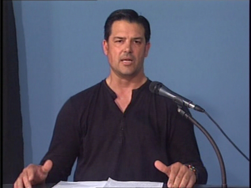 Hombre hablando en un evento público, con un fondo azul claro y un micrófono en frente de él.