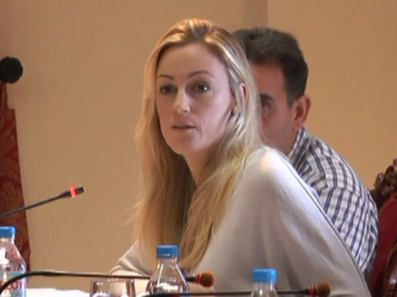Persona en una reunión o conferencia, sentada en un salón con micrófonos y botellas de agua en la mesa.