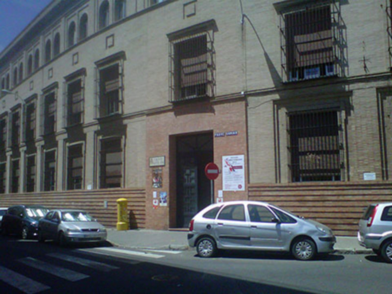 Edificio con fachada de ladrillo y ventanas con rejas, aparcamiento con coches y señal de prohibido el paso en la acera.