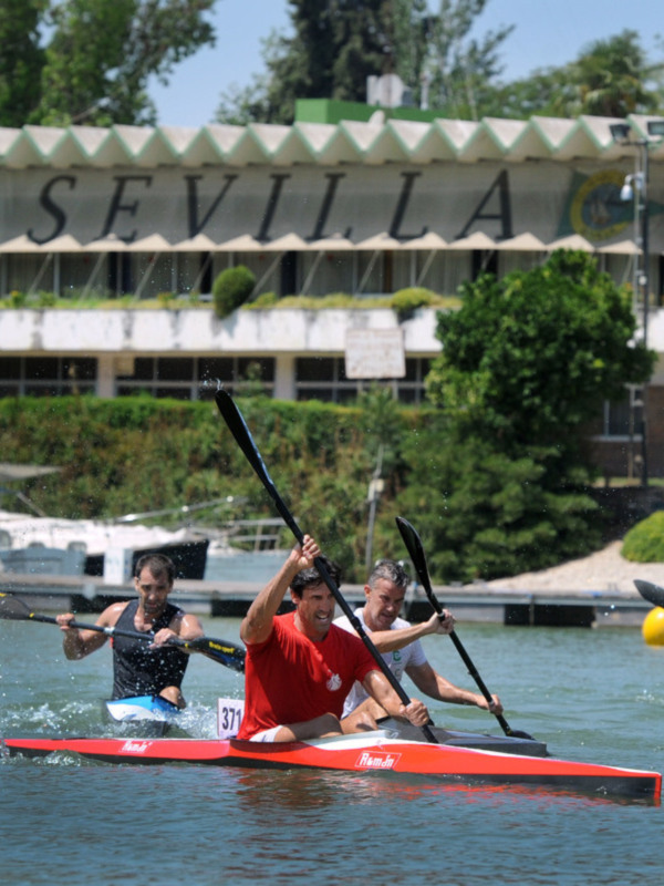 Competidores en canoa durante una carrera en el río, con el fondo de un edificio que dice "SEVILLA".
