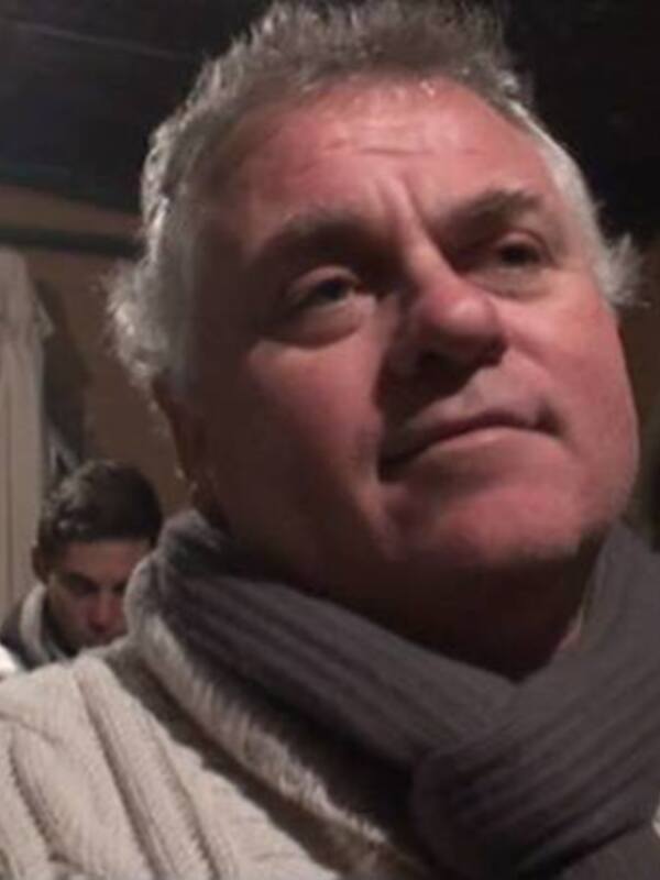 Un hombre con barba y cabello gris, mirando hacia la izquierda. Está vestido con una sudadera blanca y un suéter de lana. Fondo urbano nocturno con luces brillantes y edificios en el fondo.