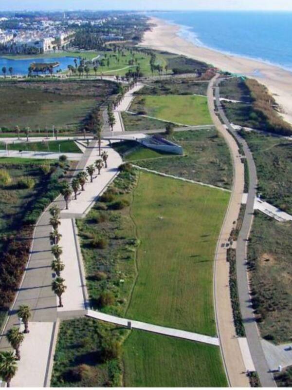 Vista aérea de un parque costero con senderos, praderas y playa, junto a edificios urbanos en el fondo.