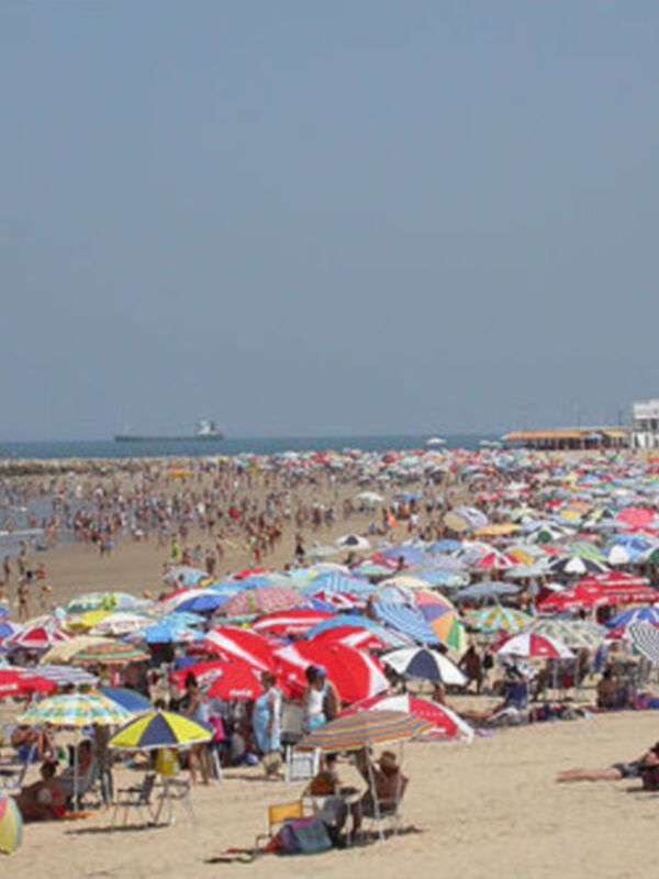 Playa con numerosas personas y sombrillas, frente a una torre de vigilancia en el horizonte.