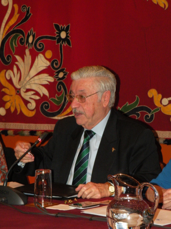 Elderly man in a suit and tie speaking at a conference, with a microphone and a red floral backdrop.