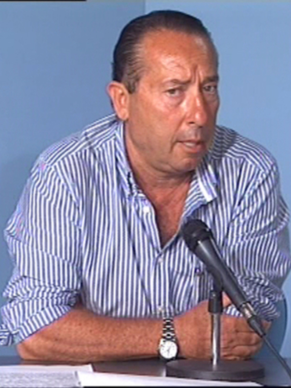 Hombre hablando en una conferencia de prensa, con micrófono y documentos en la mesa.