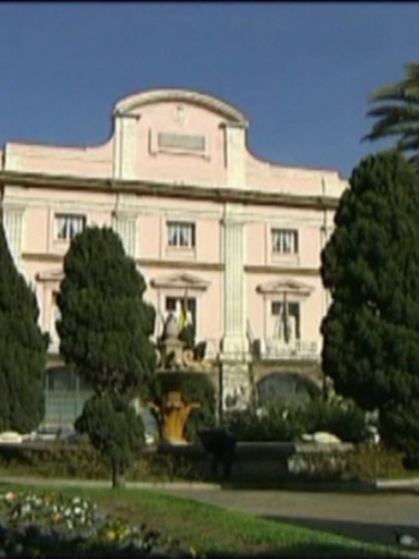 Vista frontal de un edificio neoclásico con palmeras y jardín en un día soleado.