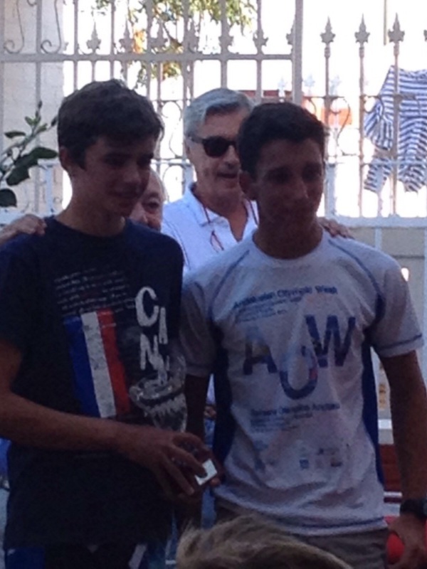 Tres personas caminando juntas, una con camiseta azul y otra con camiseta blanca, en un entorno urbano con una reja de hierro y plantas al fondo.