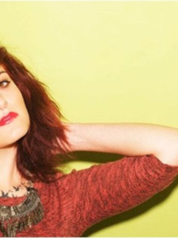 Energética mujer con cabello rojo y labios rojos, posando con brazo levantado sobre fondo verde claro.