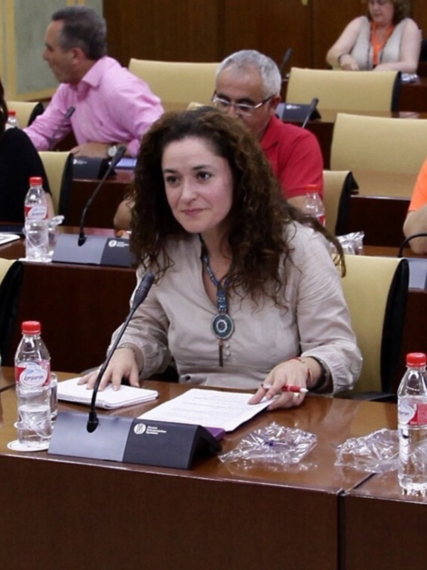 Una mujer en una sala de reuniones, sentada en una mesa con documentos y botellas de agua, con otros asistentes en el fondo.