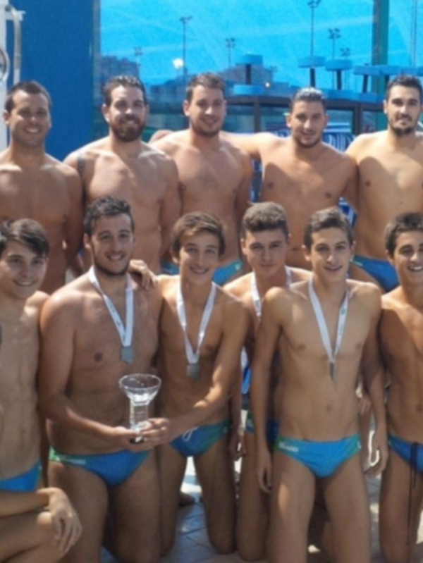 Equipo de waterpolo celebrando con medallas y trofeo en una piscina cubierta.