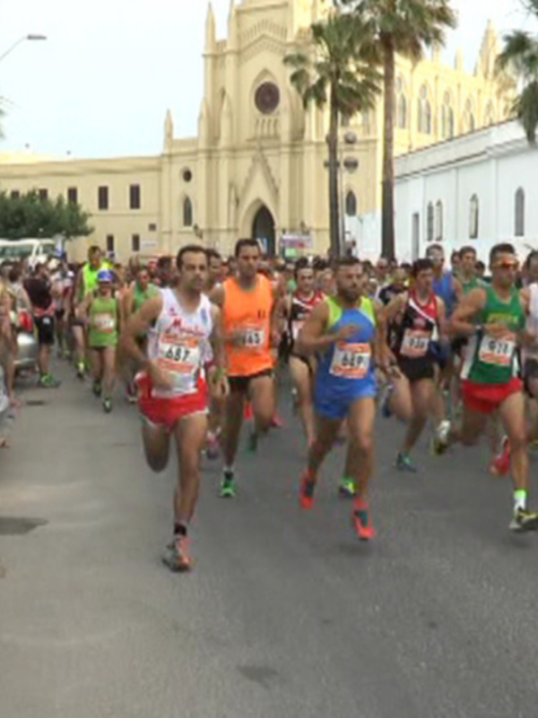 Corredores en una carrera urbana, con un edificio histórico y palmeras al fondo.