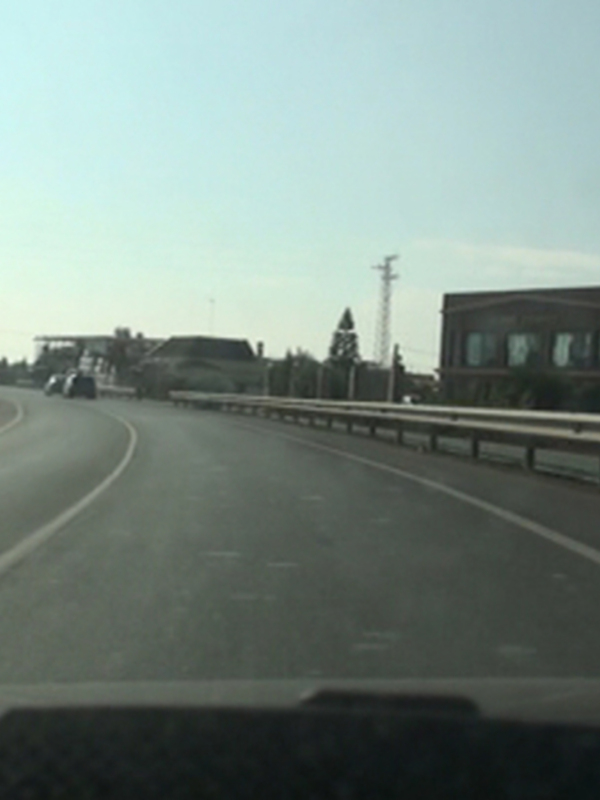 Vista desde el interior de un vehículo en una carretera recta con edificios y vegetación al costado.