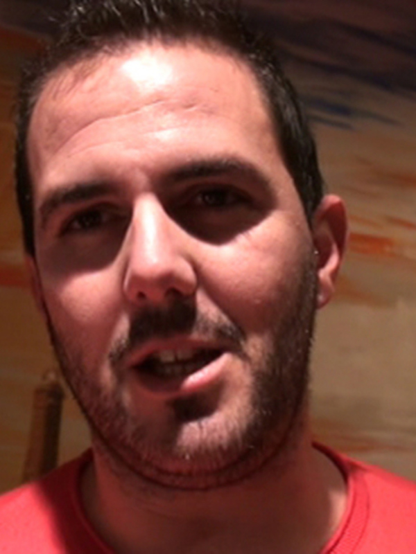 Un hombre con barba y una camiseta roja frente a una pintura de un castillo en el cielo.