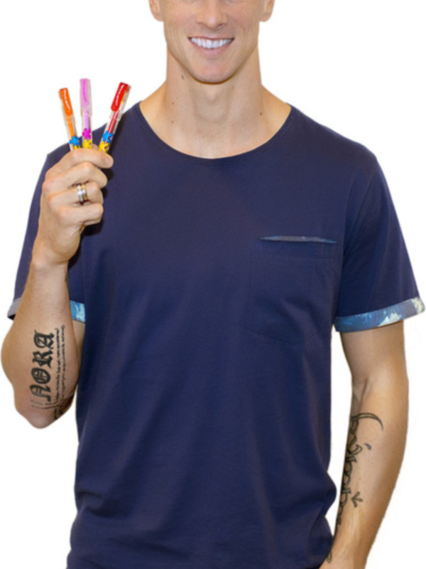 Hombre con camiseta azul y jeans, sosteniendo tres lápices de colores. Tatuajes en el brazo y una pulsera visible.