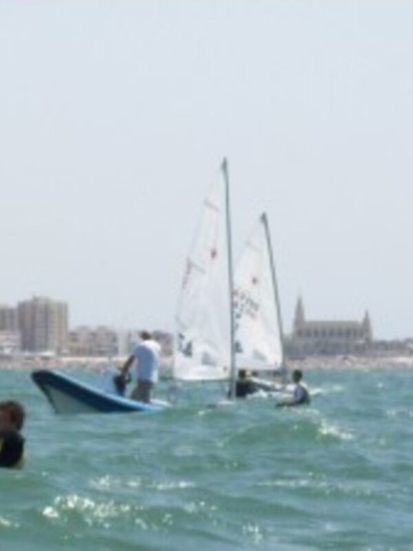 Competición de vela en el mar, con barcos navegando y personas en el agua cerca de una ciudad costera.