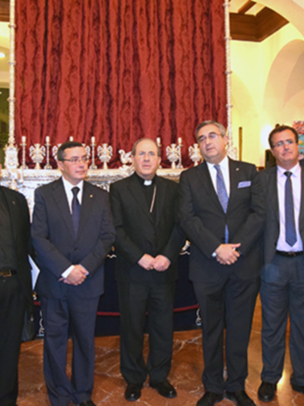 Grupo de personas en trajes formales, posando en una sala con decoración de arcos y cortinas rojas.