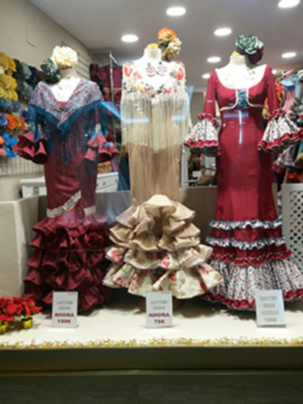 Una vitrina con tres vestidos de flamenca coloridos y volantes, junto a flores secas y etiquetas con información.
