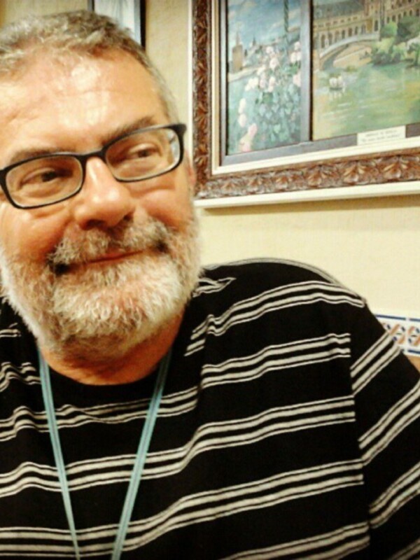 Retrato de hombre sonriente con barba y gafas, frente a un cuadro con paisaje pintado.