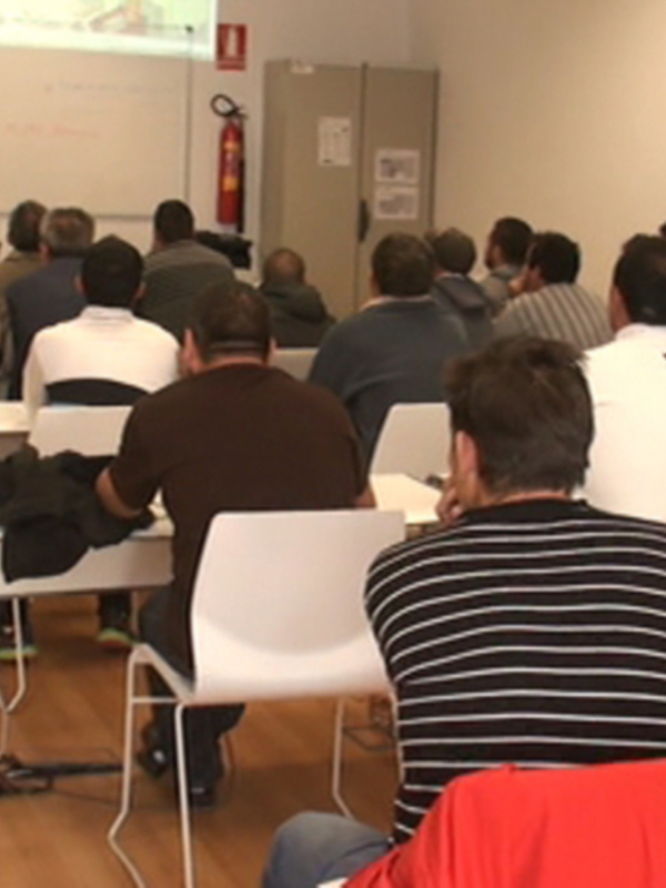 Un grupo de personas en una sala de conferencias, con una pantalla proyectando imágenes en el fondo. La sala tiene mesas blancas y sillas, y algunos participantes están escribiendo en sus cuadernos. La atmósfera parece ser de aprendizaje o presentación.