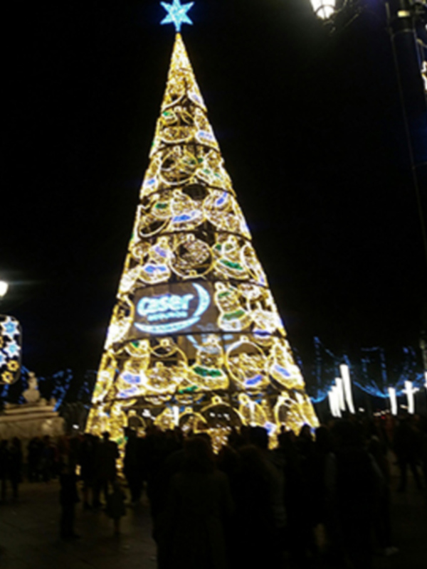 Un impresionante árbol de Navidad iluminado con luces doradas y azules, adornado con estrellas y adornos navideños, en un entorno nocturno con gente caminando alrededor.
