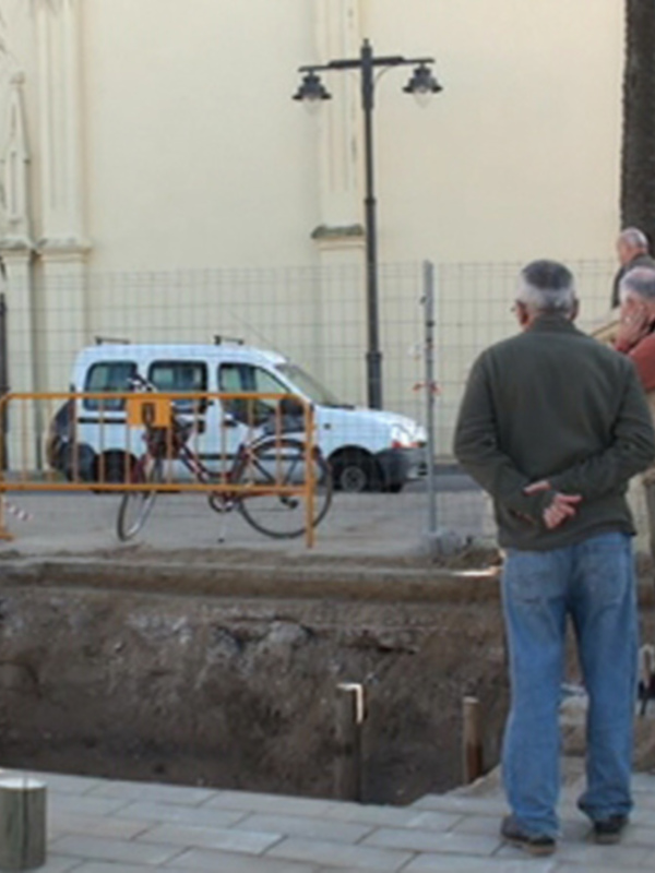 Un grupo de personas observando una excavación en el exterior de una iglesia, con un coche y una bicicleta en el fondo.
