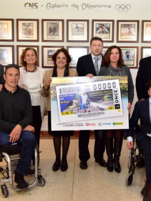 Una persona en silla de ruedas sostiene un cheque junto a otros individuos en una sala formal con banderas y fotos en la pared.