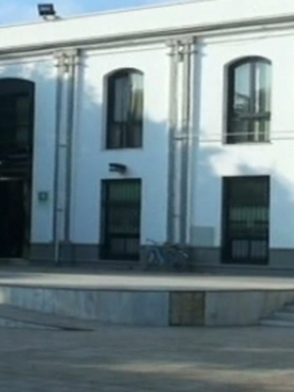 Fachada de un edificio blanco con ventanas y puertas negras, junto a un parque con árboles verdes y una persona sentada en el suelo. La imagen muestra un día soleado con cielo azul y algunas nubes dispersas.
