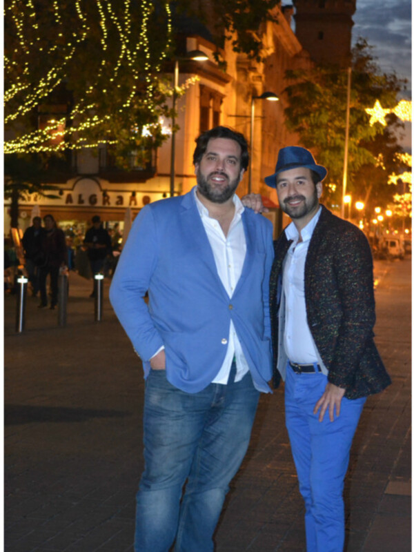 Dos hombres posan juntos en una calle nocturna adornada con luces navideñas y edificios históricos en el fondo.