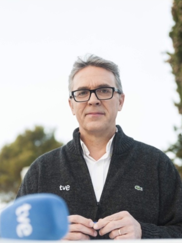 Un hombre con gafas y una chaqueta negra con un logotipo de lacoste, posando frente a un árbol en un entorno natural.