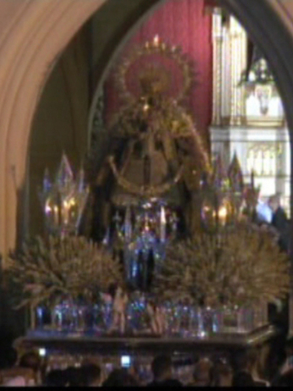 Procesión religiosa con imagen de la Virgen en un altar adornado con flores y velas, rodeada por fieles.