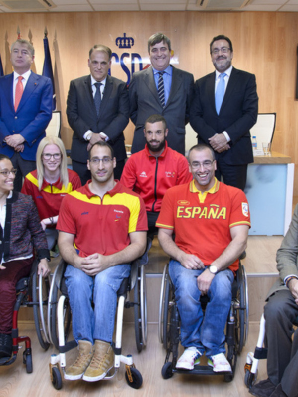 Grupo de personas en sillas de ruedas y otros miembros de la organización, posando para una foto en un entorno formal con una pantalla de televisión en el fondo.