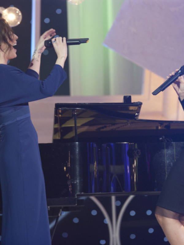 Dos mujeres cantando en escenario con piano y luces de fondo.