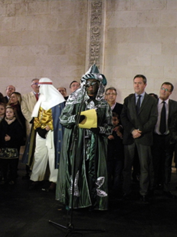 Personaje vestido de rey mago en una ceremonia pública, rodeado por espectadores en un entorno histórico nocturno.
