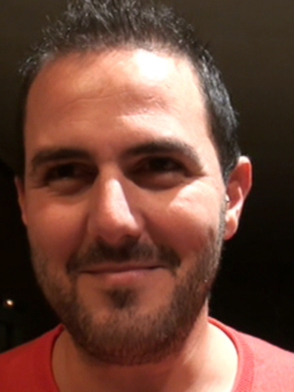 Retrato de hombre sonriente con barba, vistiendo una camiseta roja, en un ambiente interior con luces suaves.